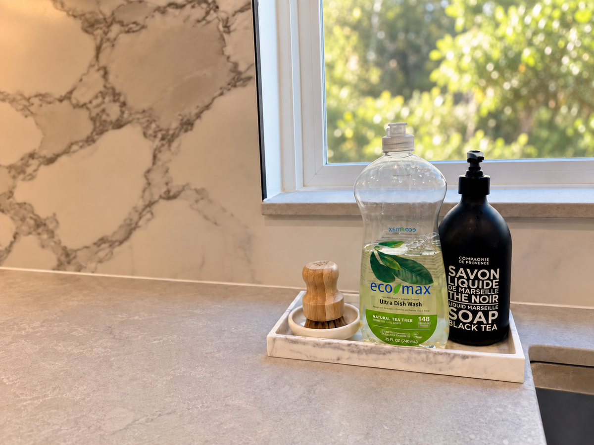 Dish soap and a wooden cleaning brush beside a kitchen window overlooking soft greenery.